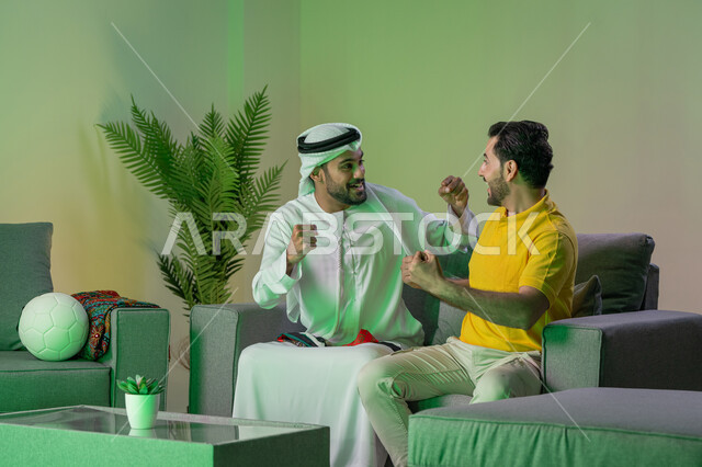 ايماءات الحماس بالفوز وتسجيل الأهداف، الشغف بمتابعة المباريات، قضاء وقت ممتع مع الأصدقاء في المنزل، صديقان عربيان خليجيان اماراتيان يشاهدان مباراة كرة القدم في غرفة المعيشة