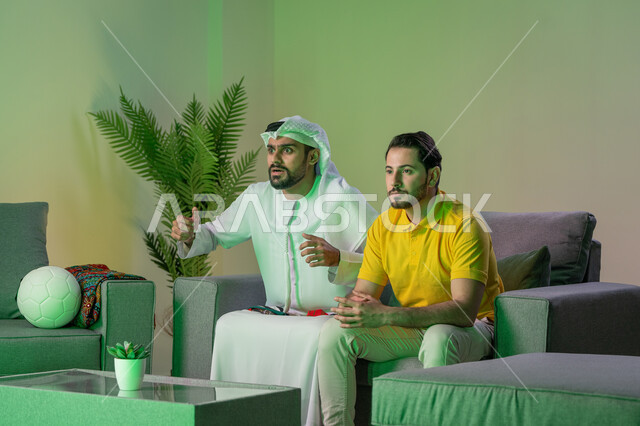 Passion for following matches, spending quality time with friends at home, two Arab Gulf and Emirati friends watching a football match in the living room, gestures of interaction and harmony with the team