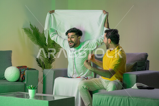 Spending free time watching TV with friends, winning and achieving goals, two young Arab Emirati men sitting on the sofa celebrating the victory of their favorite team, passion for following the football team, gestures of enthusiasm, happiness and joy