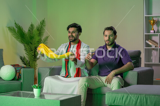متابعة و مشاهدة المباراة مع الاصدقاء, شاب عربي خليجي اماراتي يرتدي الكندورة البيضاء يمسك بيده الوشاح الاصفر يجلس مع صديقه في غرفة المعيشة يشاهدان مباراة كرة القدم، قضاء وقت ممتع مع الاصدقاء, ايماءات المنافسة والتحدي , أندية وتشجيع .