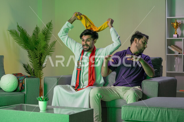 التحدي مع الاصدقاء و مشاهدة المباراة في المنزل ,صديقان عربيان اماراتيان خليجيان يشاهدان مباراة للدوري العربي ، شاب يشجع فريق و صديقه يشجع خصمه , ايماءات الحماس و المنافسة , أندية وتشجيع .