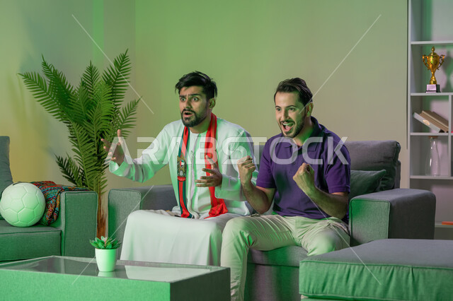 Watching matches at home and challenging with friends, two Gulf Arab Emirati friends watching an Arab League match, gestures of competition and challenge, clubs and encouragement.
