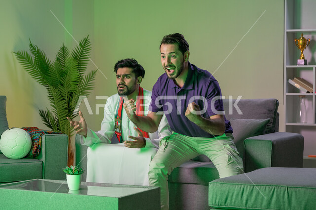 Watching the match and being excited with friends, two young Arab Emirati Gulf men watching an Arab League match at home, gestures of enthusiasm and challenge, clubs and encouragement.