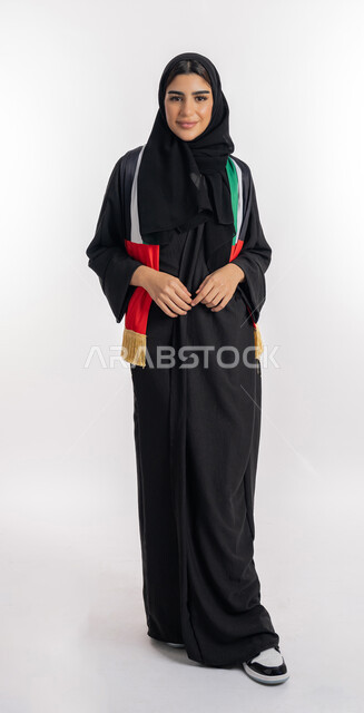الوقوف باستقامة والنظر إلى الكاميرا بإيماءات البهجة والسرور، مشجعة للفرق والنوادي الوطنية، اهتمام الفتيات بمتابعة المباريات، بورتريه لشابة عربية خليجية إماراتيه ترتدي العباية والحجاب ووشاح تشجيع يحمل علم الإمارات، صورة لكامل الجسم، خلفية بيضاء