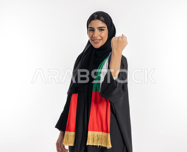 Looking at the camera with joyful gestures, girls’ interest in watching sports matches, a Saudi girl wearing a cheer scarf bearing the UAE flag, a fan of national teams and clubs, a portrait of an Arab Gulf Emirati young woman wearing an abaya and hijab, raising her hand with victory gestures, white background.