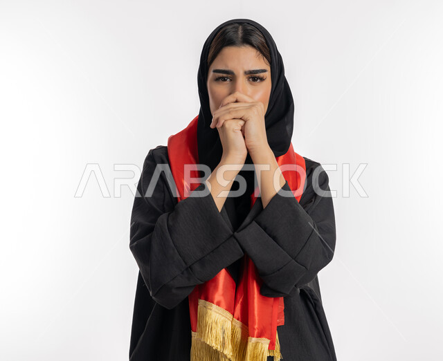 النظر إلى الكاميرا بتعابير التوتر، مشجعة للفرق الوطنية، اهتمام الفتيات بمتابعة المباريات، مفهوم تشجيع الفريق المفضل، بورتريه لشابة عربية خليجية إماراتيه ترتدي العباية والحجاب ووشاح باللون الأحمر، فتاة سعودية تقوم بشبك أيديها أمام فمها بإيماءات الخوف، خلفية بيضاء