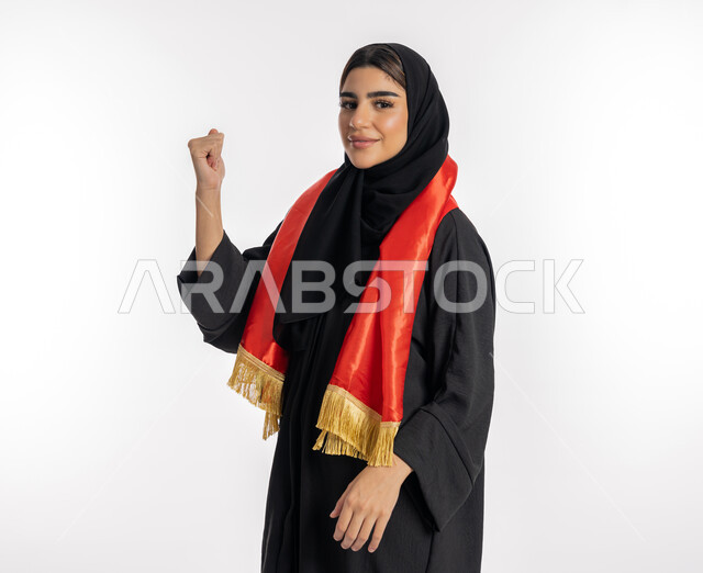 مشجعة للفرق الوطنية، النظر إلى الكاميرا بتعابير التحدي، اهتمام الفتيات بمتابعة المباريات، مفهوم تشجيع الفريق المفضل، بورتريه لشابة عربية خليجية إماراتيه ترتدي العباية والحجاب ووشاح باللون الأحمر، فتاة سعودية تقوم برفع يديها بإيماءات النصر، خلفية بيضاء