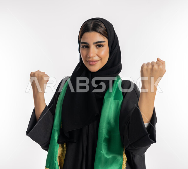 تشجيع للفرق الوطنية، النظر إلى الكاميرا بتعابير التحدي والقوة، اهتمام الفتيات بمتابعة المباريات، مفهوم تشجيع الفريق المفضل، بورتريه لشابة عربية خليجية إماراتيه ترتدي العباية والحجاب ووشاح باللون الأخضر، فتاة سعودية تقوم برفع يديها بإيماءات النصر، خلفية بيضاء