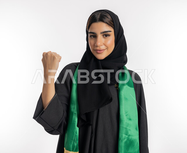 النظر إلى الكاميرا بتعابير التحدي والقوة، مشجعة للفرق الوطنية، اهتمام الفتيات بمتابعة المباريات، مفهوم تشجيع الفريق المفضل، بورتريه لشابة عربية خليجية إماراتيه ترتدي العباية والحجاب ووشاح باللون الأخضر، فتاة سعودية تقوم برفع يدها بإيماءات النصر، خلفية بيضاء