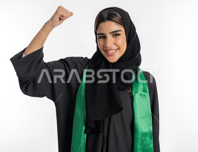 فتاة سعودية تنظر إلى الكاميرا بتعابير الفرح، مشجعة للفرق والنوادي الوطنية، اهتمام الفتيات بمتابعة المباريات، مفهوم تشجيع النادي المفضل، بورتريه لشابة عربية خليجية إماراتيه ترتدي العباية والحجاب ووشاح باللون الأخضر تقوم برفع يدها بإيماءات النصر، خلفية بيضاء