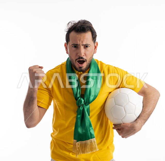 إيماءات يدين تدل على الحماس والقوة، الشغف في متابعة المباريات الرياضية، النظر إلى الكاميرا بتعابير الدهشة والذهول، مشجع للفرق والنوادي الوطنية، بورتريه لشاب عربي خليجي إماراتي يرتدي الزي الكاجوال ووشاح تشجيع باللون الأخضر يقوم بحمل كرة باللون الأبيض، خلفية بيضاء