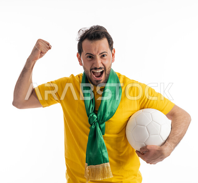 A supporter of national teams and clubs, hand gestures indicating enthusiasm and strength, passion for following sports matches, looking at the camera with expressions of astonishment and astonishment, portrait of a young Arab Gulf Emirati man wearing a casual outfit and a green cheerleading scarf, carrying a white ball, white background.