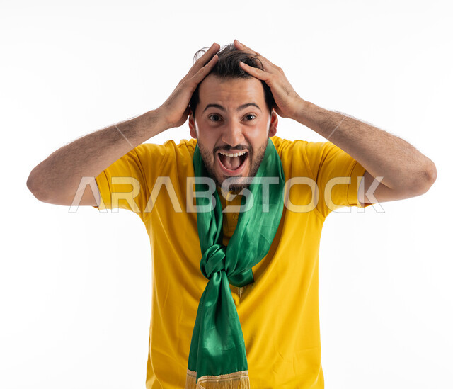 النظر إلى الكاميرا بإيماءات الدهشة والذهول، الشغف في متابعة المباريات الرياضية، مشجع للفرق والنوادي الوطنية، بورتريه لشاب عربي خليجي إماراتي يرتدي الزي الكاجوال ووشاح تشجيع باللون الأخضر يضع يديه على رأسه بتعابير الحماس، خلفية بيضاء