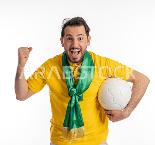 إيماءات يدين تدل على الحماس والقوة، الشغف في متابعة المباريات الرياضية، النظر إلى الكاميرا بتعابير الدهشة والذهول، مشجع للفرق والنوادي الوطنية، بورتريه لشاب عربي خليجي إماراتي يرتدي الزي الكاجوال ووشاح تشجيع باللون الأخضر يقوم بحمل كرة باللون الأبيض، خلفية بيضاء