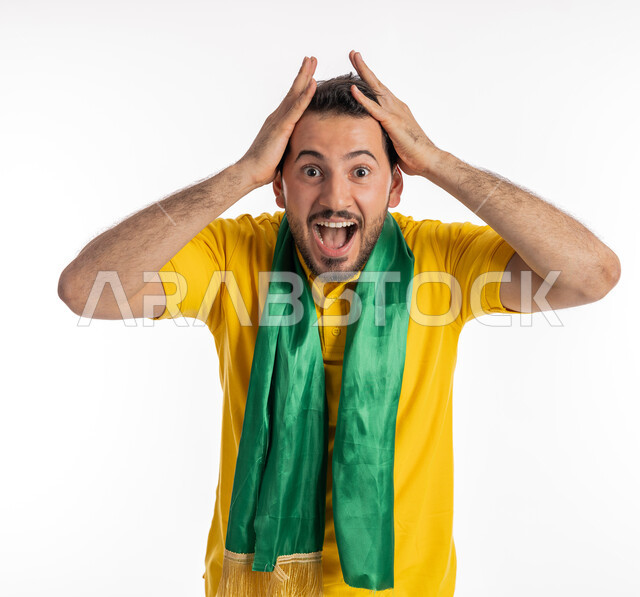 النظر إلى الكاميرا بإيماءات الدهشة والذهول، الشغف في متابعة المباريات الرياضية، مشجع للفرق والنوادي الوطنية، بورتريه لشاب عربي خليجي إماراتي يرتدي الزي الكاجوال ووشاح تشجيع باللون الأخضر يضع يديه على رأسه بتعابير الحماس، خلفية بيضاء