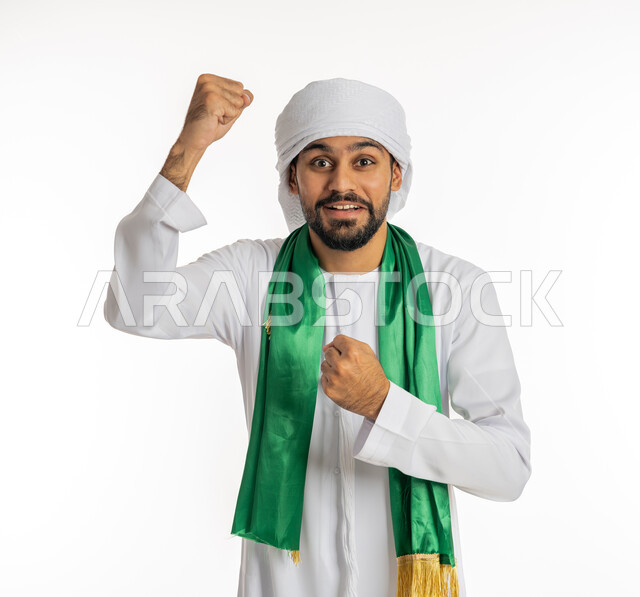 النظر إلى الكاميرا بتعابير وجه تدل على الحماس والشغف، إيماءات يدين تدل على النصر، مشجع للفرق والنوادي الرياضية الإماراتية، بورتريه لشاب عربي خليجي إماراتي يرتدي الكندورة مع العمامة ووشاح تشجيع باللون الأخضر، خلفية بيضاء