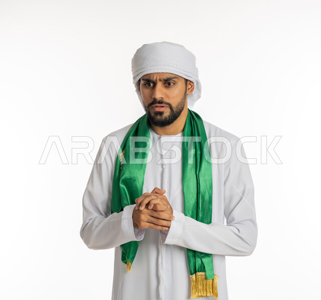 شبك الأيدي ببعضها بإيماءات التوتر والتفكير بشيء ما، الشغف في متابعة المباريات الرياضية، مشجع للفريق الأهلي والنوادي الوطنية، بورتريه لشاب عربي خليجي إماراتي يرتدي الكندورة الإمارتية مع العمامة ووشاح تشجيع باللون الأخضر