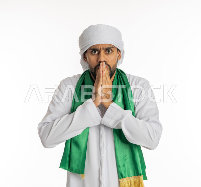 النظر إلى الكاميرا بتعابير الغضب، الشغف في متابعة المباريات الرياضية، مشجع للفريق الأهلي والنوادي الوطنية، بورتريه لشاب عربي خليجي إماراتي يرتدي الكندورة الإمارتية مع العمامة ووشاح تشجيع باللون الأخضر يضع يديه أمام فمه بإيماءات التوتر