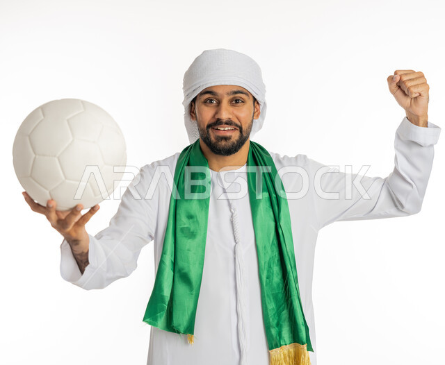 رفع الكرة بإيماءات النصر، النظر إلى الكاميرا بتعابير وجه تدل على الحماس والشغف، إيماءة يد تدل على القوة والتحدي، مشجع للفرق والنوادي الرياضية الإماراتية، بورتريه لشاب عربي خليجي إماراتي يرتدي الكندورة مع العمامة ووشاح تشجيع باللون الأخضر، خلفية بيضاء
