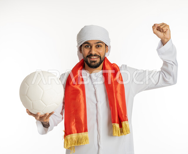 رفع الكرة بإيماءات النصر، النظر إلى الكاميرا بتعابير وجه تدل على الحماس والشغف، إيماءة يد تدل على القوة والتحدي، مشجع للفرق والنوادي الرياضية الإماراتية، بورتريه لشاب عربي خليجي إماراتي يرتدي الكندورة مع العمامة ووشاح تشجيع باللون الأحمر، خلفية بيضاء