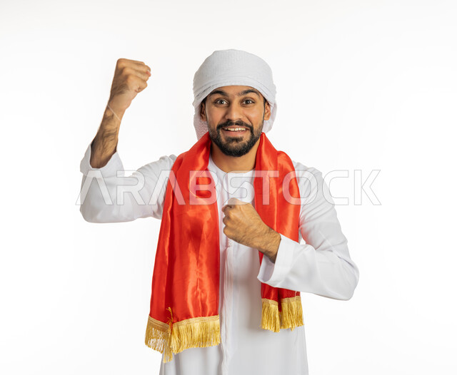 النظر إلى الكاميرا بتعابير وجه تدل على الحماس والشغف، إيماءة يد تدل على النصر، مشجع للفرق والنوادي الرياضية الإماراتية، بورتريه لشاب عربي خليجي إماراتي يرتدي الكندورة مع العمامة ووشاح تشجيع باللون الأحمر، خلفية بيضاء