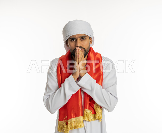 النظر إلى الكاميرا بتعابير الغضب، الشغف في متابعة المباريات الرياضية، مشجع للفريق الأهلي والنوادي الوطنية، بورتريه لشاب عربي خليجي إماراتي يرتدي الكندورة الإمارتية مع العمامة ووشاح تشجيع باللون الأحمر يضع يديه أمام فمه بإيماءات التوتر، خلفية بيضاء