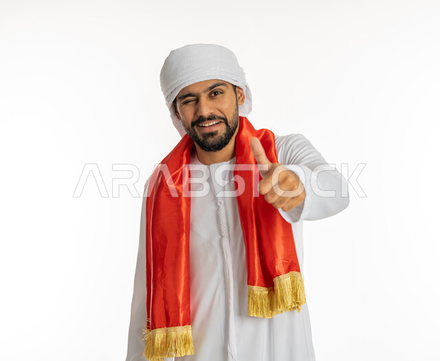ايماءات وجه ويدين تدل على التحدي، مشجع للفريق الأهلي والنوادي الوطنية، الشغف في متابعة المباريات الرياضية، النظر إلى الكاميرا بتعابير السرور، بورتريه مقرب لشاب عربي خليجي إماراتي يرتدي الكندورة الإمارتية مع العمامة ووشاح تشجيع باللون الأحمر، خلفية بيضاء
