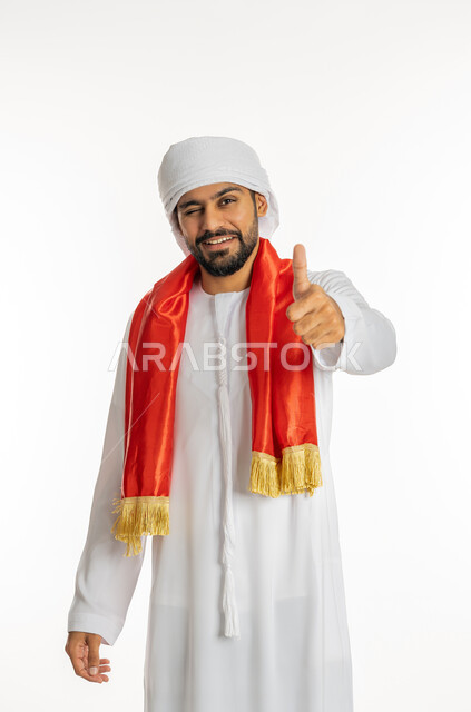 النظر إلى الكاميرا بتعابير السرور، ايماءات وجه ويدين تدل على التحدي، مشجع للفريق الأهلي والنوادي الوطنية، الشغف في متابعة المباريات الرياضية، بورتريه مقرب لشاب عربي خليجي إماراتي يرتدي الكندورة الإمارتية مع العمامة ووشاح تشجيع باللون الأحمر، خلفية بيضاء