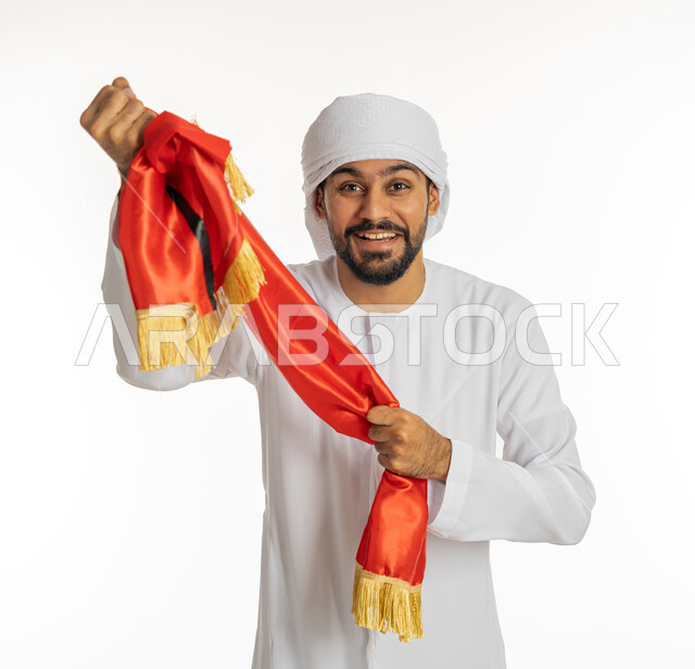 الإمساك بوشاح تشجيع باللون الأحمر والنظر إلى الكاميرا بإيماءات الفوز، الشغف في متابعة المباريات الرياضية، مشجع للفريق الأهلي والنوادي الوطنية، بورتريه لشاب عربي خليجي إماراتي يرتدي الكندورة الإمارتية مع العمامة، خلفية بيضاء