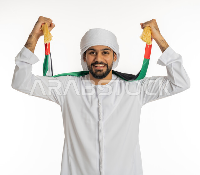 إيماءات الحماس والنصر، النظر إلى الكاميرا بتعابير البهجة والسرور، مشجع للفرق والنوادي الرياضية الإماراتية، بورتريه لشاب عربي خليجي إماراتي يرتدي الكندورة مع العمامة ووشاح تشجيع يحمل علم الإمارات العربية المتحدة يقوم بإمساكه بيديه، خلفية بيضاء