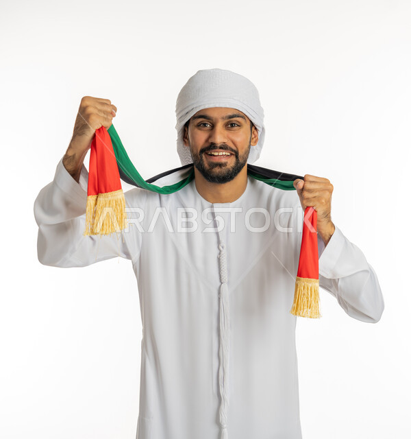 تعابير تدل على النصر والفوز، النظر إلى الكاميرا بإيماءات البهجة والسرور، مشجع للفرق والنوادي الرياضية الإماراتية، بورتريه لشاب عربي خليجي إماراتي يرتدي الكندورة مع العمامة ووشاح تشجيع يحمل علم الإمارات العربية المتحدة يقوم بإمساكه بيديه، خلفية بيضاء