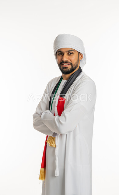 النظر إلى الكاميرا بإيماءات البهجة والسرور، مشجع للفرق والنوادي الرياضية الإماراتية، بورتريه لشاب عربي خليجي إماراتي يرتدي الكندورة مع العمامة ووشاح تشجيع يحمل علم الإمارات العربية المتحدة يقف بأيدي مكتوفة، خلفية بيضاء