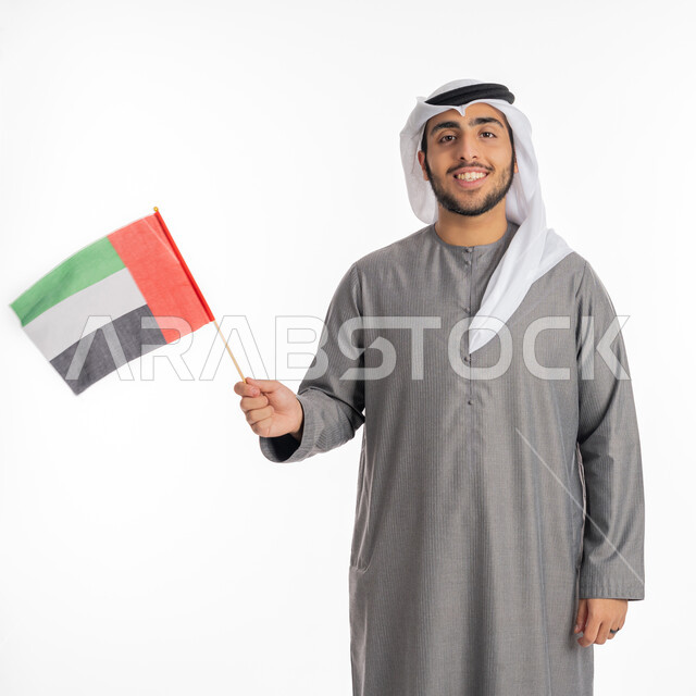 ذكرى يوم الاتحاد والاستقلال الإماراتي 2 ديسمبر، صورة لشاب إماراتي عربي خليجي مبتسم يرتدي الزي التقليدي ينظر إلى الكاميرا ويحمل بيده علم دولة الإمارات، الاحتفال بيوم العلم الإماراتي 3 نوفمبر، الأعياد والمناسبات الوطنية، إيماءات الشموخ والاعتزاز بالانتماء للوطن العربي