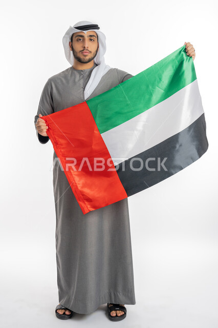 اليوم الوطني لاتحاد الإمارات العربية 2 ديسمبر، بورتريه لشاب إماراتي عربي خليجي يرتدي الزي التقليدي ينظر إلى الكاميرا ويحمل بيديه علم الإمارات، إيماءات الفخر والاعتزاز بالانتماء للوطن العربي، الاحتفال بيوم العلم الإماراتي 3 نوفمبر، خلفية بيضاء، الطول الكامل للجسم
