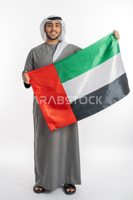 اليوم الوطني لاستقلال الإمارات 2 ديسمبر، بورتريه لشاب إماراتي عربي خليجي مبتسم يرتدي الزي التقليدي ينظر إلى الكاميرا ويحمل بيده علم دولة الإمارات، إيماءات الفخر والاعتزاز بالانتماء للوطن، الاحتفال بيوم العلم الإماراتي 3 نوفمبر، خلفية بيضاء، الطول الكامل للجسم