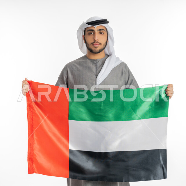 اليوم الوطني لاتحاد الإمارات العربية 2 ديسمبر، بورتريه لشاب إماراتي عربي خليجي يرتدي الزي التقليدي ينظر إلى الكاميرا ويحمل بيديه علم دولة الإمارات، إيماءات الفخر والاعتزاز بالانتماء للوطن العربي، الاحتفال بيوم العلم الإماراتي 3 نوفمبر، خلفية بيضاء