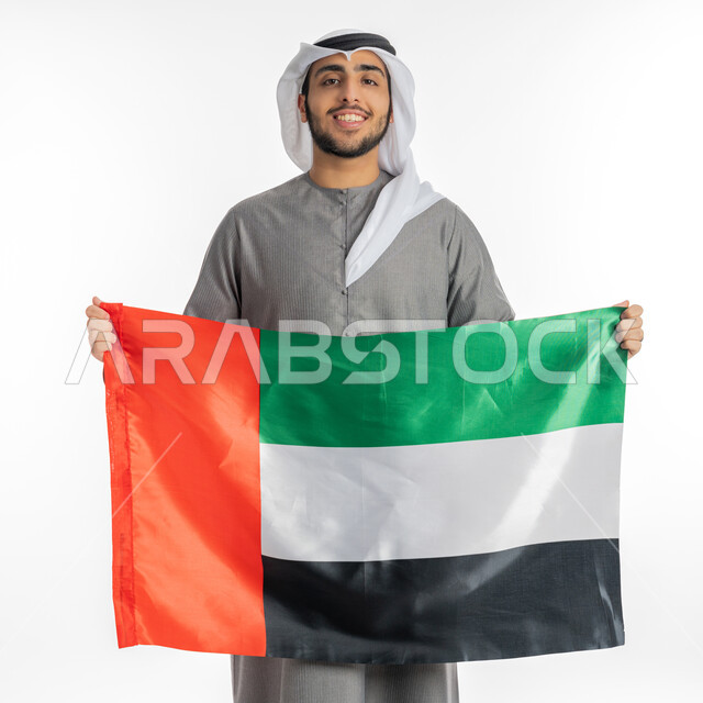 اليوم الوطني لاستقلال واتحاد الإمارات 2 ديسمبر، بورتريه لشاب إماراتي عربي خليجي يرتدي الزي التقليدي مبتسم ينظر إلى الكاميرا ويحمل بيديه علم دولة الإمارات، إيماءات الفخر والاعتزاز بالانتماء للوطن العربي، الاحتفال بيوم العلم الإماراتي 3 نوفمبر، خلفية بيضاء