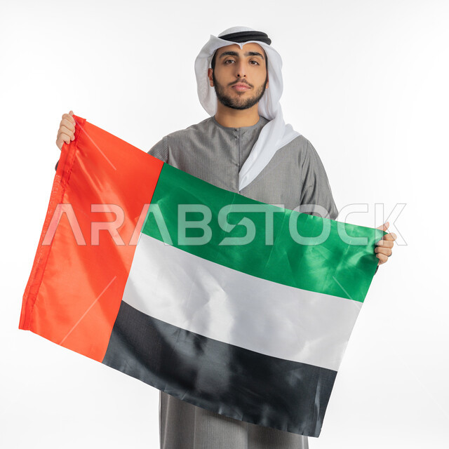 الاحتفال باليوم الوطني لاستقلال الإمارات 2 ديسمبر، بورتريه لشاب إماراتي عربي خليجي يرتدي الزي التقليدي ينظر إلى الكاميرا ويحمل بيديه علم دولة الإمارات، إيماءات الشموخ والفخر والاعتزاز بالانتماء للوطن العربي، الاحتفال بيوم العلم الإماراتي 3 نوفمبر، خلفية بيضاء