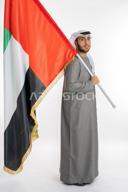 UAE National Independence Day, December 2, portrait of a young Emirati Arab Gulf man wearing traditional clothing looking at the camera and holding the UAE flag in his hand, gestures of pride and pride in belonging to the Arab world, celebrating UAE Flag Day November 3, white background, full length of the body
