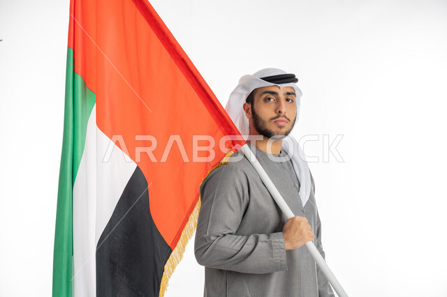 اليوم الوطني لاستقلال الإمارات 2 ديسمبر، بورتريه لشاب إماراتي عربي خليجي يرتدي الزي التقليدي ينظر إلى الكاميرا ويحمل بيده علم دولة الإمارات، إيماءات الفخر والاعتزاز بالانتماء للوطن العربي، الاحتفال بيوم العلم الإماراتي 3 نوفمبر، خلفية بيضاء