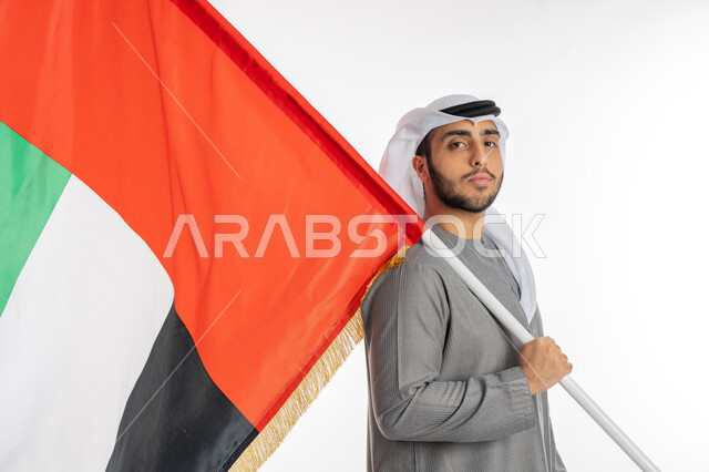 ذكرى يوم الاستقلال الإماراتي 2 ديسمبر، بورتريه لشاب إماراتي عربي خليجي يرتدي الزي التقليدي ينظر إلى الكاميرا ويحمل بيده علم دولة الإمارات، إيماءات الشموخ والإباء والاعتزاز بالانتماء للوطن العربي، الاحتفال بيوم العلم الإماراتي 3 نوفمبر، خلفية بيضاء