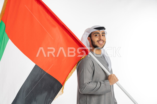 المناسبات الوطنية الإماراتية، يوم الاستقلال والاتحاد الإماراتي 2 ديسمبر، بورتريه لشاب إماراتي عربي خليجي مبتسم يرتدي الزي التقليدي ينظر إلى الكاميرا ويحمل بيده علم الإمارات، إيماءات الاحترام والفرح بالانتماء للوطن، الاحتفال بيوم العلم الإماراتي 3 نوفمبر، خلفية بيضاء