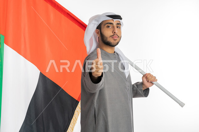 يوم الاستقلال والاتحاد الإماراتي 2 ديسمبر، بورتريه لشاب إماراتي عربي خليجي يرتدي الزي التقليدي ينظر إلى الكاميرا ويحمل بيده علم دولة الإمارات، إيماءات الافتخار والشموخ بالانتماء للوطن، الاحتفال بيوم العلم الإماراتي 3 نوفمبر، الأعياد والمناسبات الوطنية، خلفية بيضاء