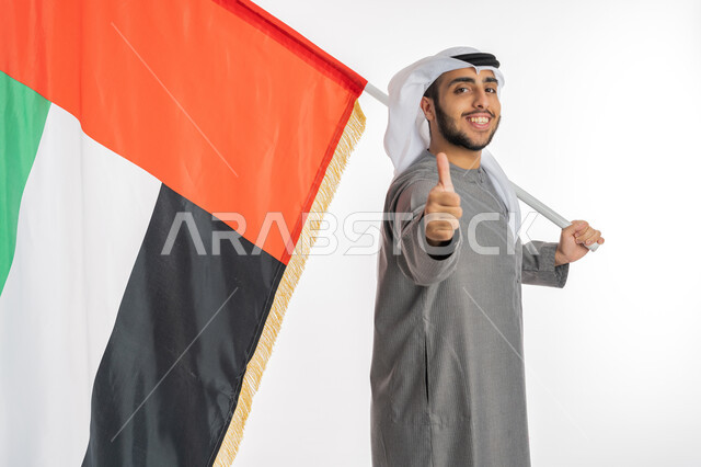 Commemoration of UAE Independence Day, December 2, portrait of a young Emirati Arab Gulf man wearing traditional clothing, smiling, looking at the camera and holding the UAE flag in his hand, gestures of admiration, pride, and pride in belonging to the Arab world, celebrating UAE Flag Day, November 3, white background