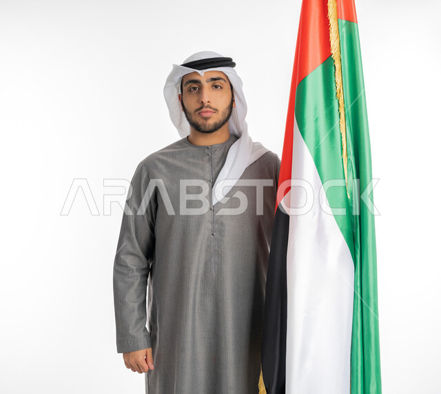 الاحتفال بيوم الاستقلال الإماراتي 2 ديسمبر، بورتريه لشاب إماراتي عربي خليجي يرتدي الزي التقليدي ينظر إلى الكاميرا ويقف بجانب علم دولة الإمارات، علامات الفخر والاعتزاز بالانتماء للوطن العربي، الاحتفال بيوم العلم الإماراتي 3 نوفمبر، خلفية بيضاء