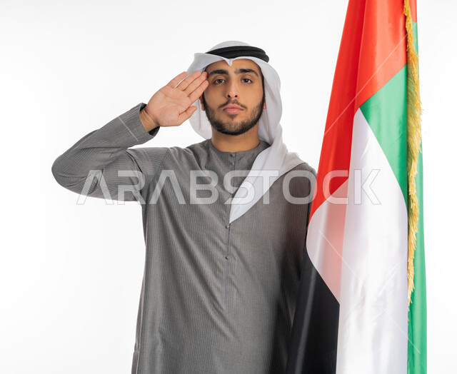 يوم الاستقلال الإماراتي 2 ديسمبر، بورتريه لشاب إماراتي عربي خليجي يرتدي الزي التقليدي ينظر إلى الكاميرا ويقف بجانب علم دولة الإمارات، إيماءات القاء التحية والاحترام للعلم الإماراتي، الاحتفال بيوم العلم الإماراتي 3 نوفمبر، المناسبات والأعياد الوطنية، خلفية بيضاء