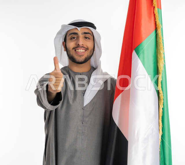 ذكرى الاتحاد الإماراتي 2 ديسمبر، بورتريه لشاب إماراتي عربي خليجي مبتسم يرتدي الزي التقليدي ينظر إلى الكاميرا ويقف بجانب علم دولة الإمارات، إيماءات الإعجاب والفرح بالإنتماء للوطن العربي، الاحتفال بيوم العلم الإماراتي 3 نوفمبر، المناسبات والأعياد الوطنية، خلفية بيضاء