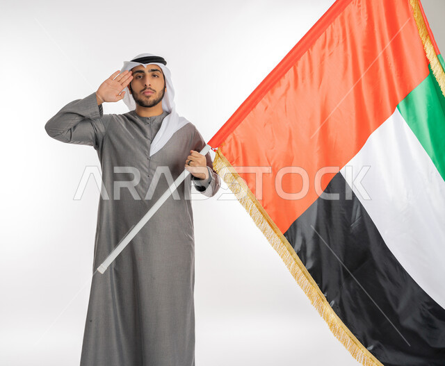 ذكرى يوم الاستقلال الإماراتي 2 ديسمبر، بورتريه لشاب إماراتي عربي خليجي يرتدي الزي التقليدي ينظر إلى الكاميرا ويحمل بيده علم دولة الإمارات، إيماءات القاء التحية والاحترام للعلم الإماراتي، الاحتفال بيوم العلم الإماراتي 3 نوفمبر، الأعياد والمناسبات الوطنية، خلفية بيضاء