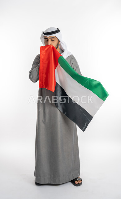 Commemoration of UAE Union and Independence Day, December 2, portrait of a young Emirati Arab Gulf man wearing traditional clothing holding the UAE flag in his hand and kissing it, celebrating UAE Flag Day, November 3, gestures of gratitude and admiration for belonging to the Arab world, white background, full length of the body