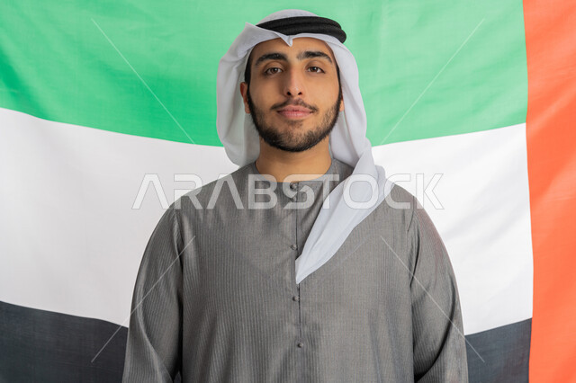 ذكرى اليوم الوطني الإماراتي للاستقلال 2 ديسمبر، صورة لشاب إماراتي عربي خليجي يرتدي الزي التقليدي يقف بشموخ وينظر إلى الكاميرا بايماءات الحب والفرح بالانتماء للوطن وخلفه العلم الإماراتي، الاحتفال بيوم العلم الإماراتي 3 نوفمبر، الأعياد والمناسبات الوطنية